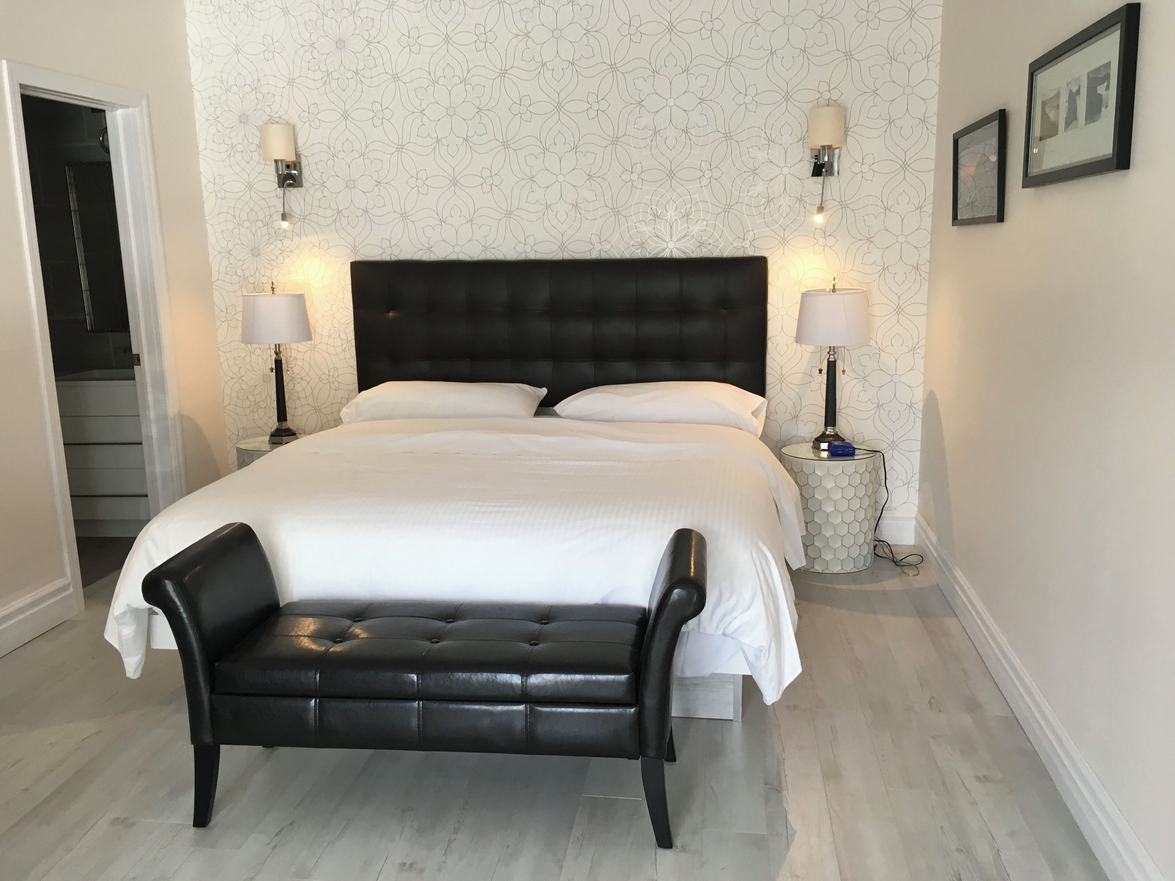A hotel bedroom with a large bed, two bedside lamps, a black leather bench, and a patterned wallpaper.