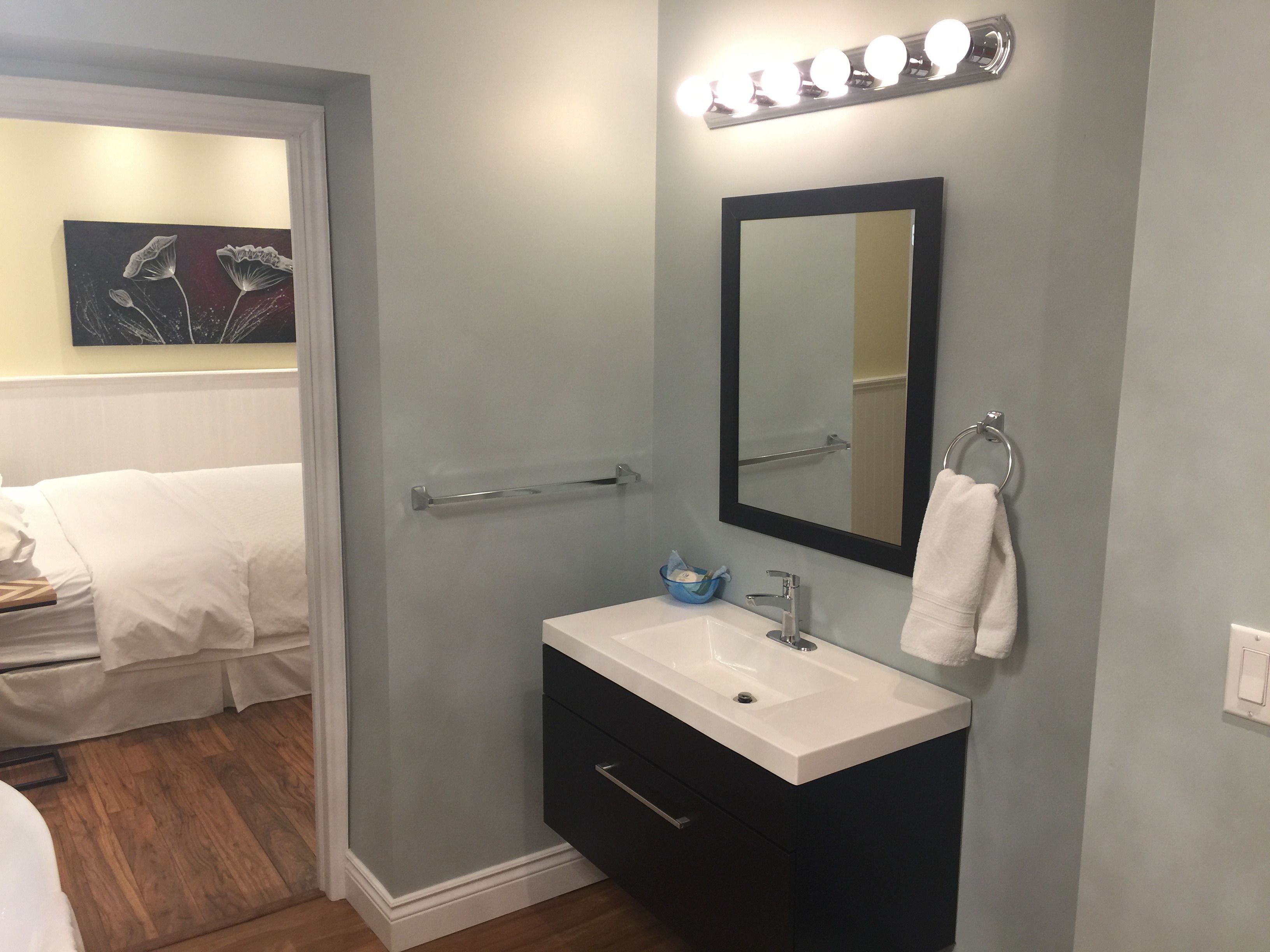 Un baño con un lavamanos blanco, un espejo grande y un perchero. Hay un cuadro en la pared y una puerta que da a otra habitación.