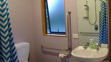 A bathroom with a white toilet, a window, a shower with a grab bar, and a sink with soap dispenser.