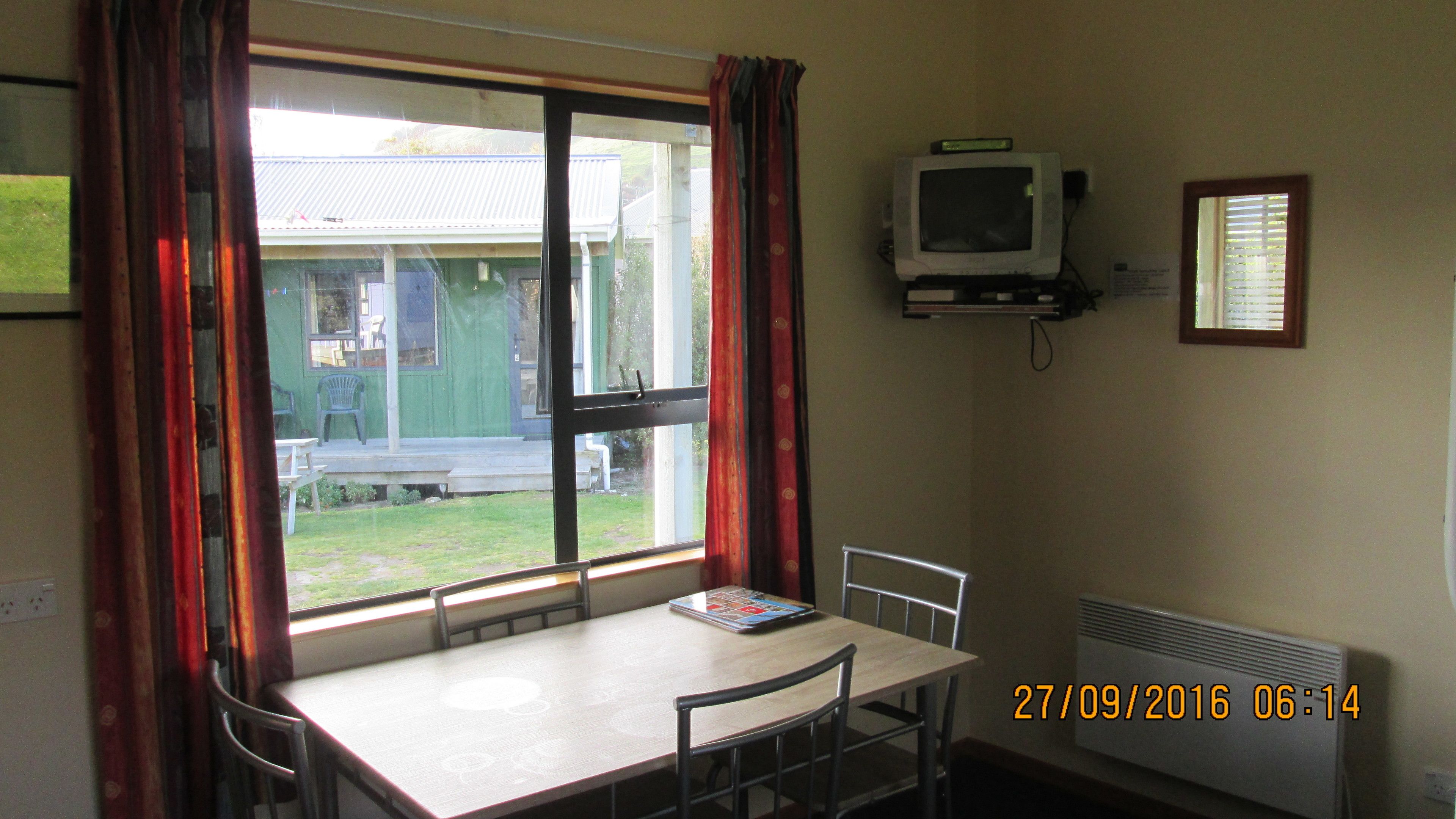 Ein Zimmer mit Tisch, Stühlen, einem Fenster mit Vorhängen, einem Fernseher und einem Heizkörper.