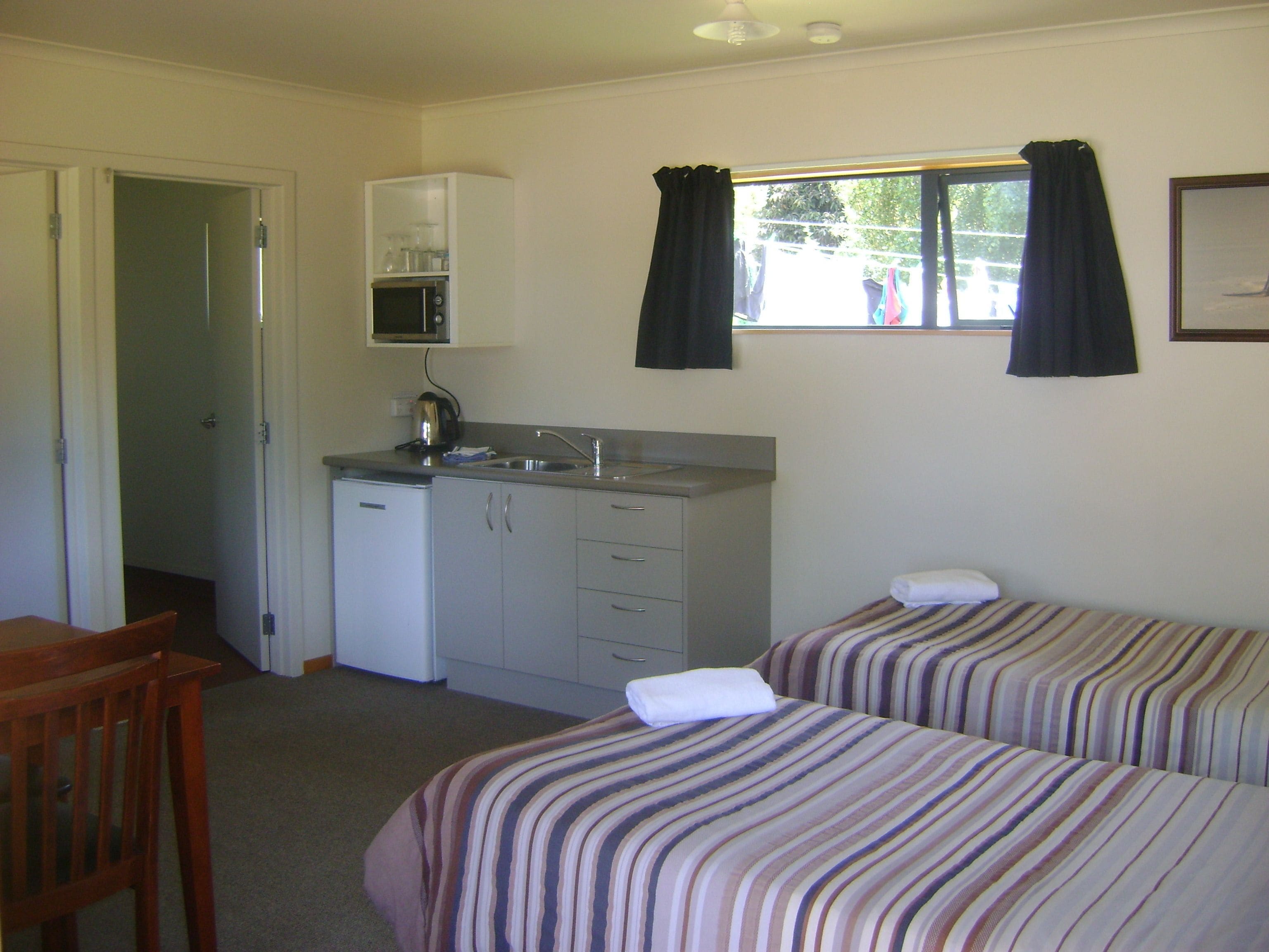 Une chambre compacte avec deux lits simples, une petite kitchenette équipée d’un micro-ondes et une fenêtre avec des rideaux.