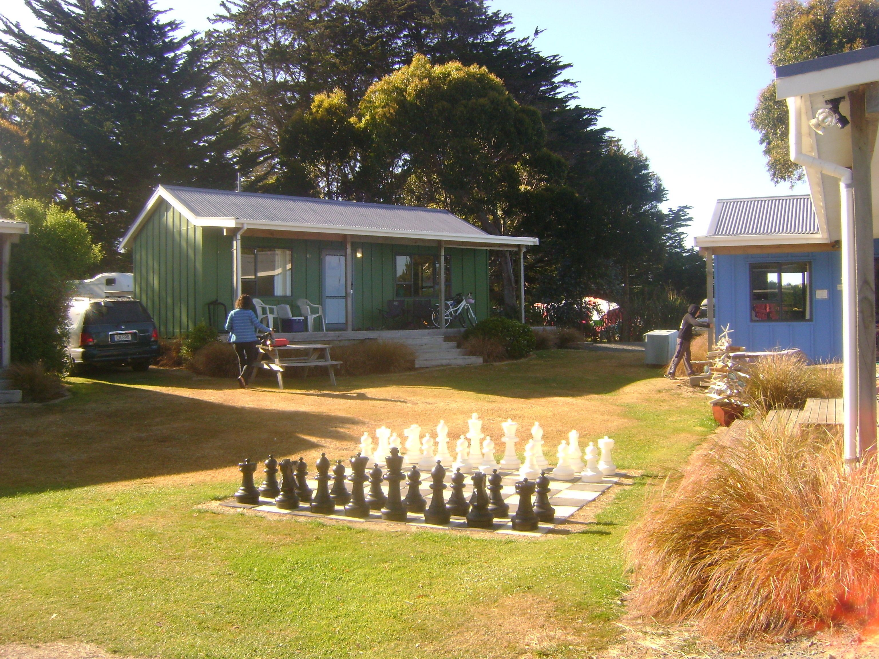 A residential area with a large outdoor chessboard, a small house with a porch, and a car parked nearby.