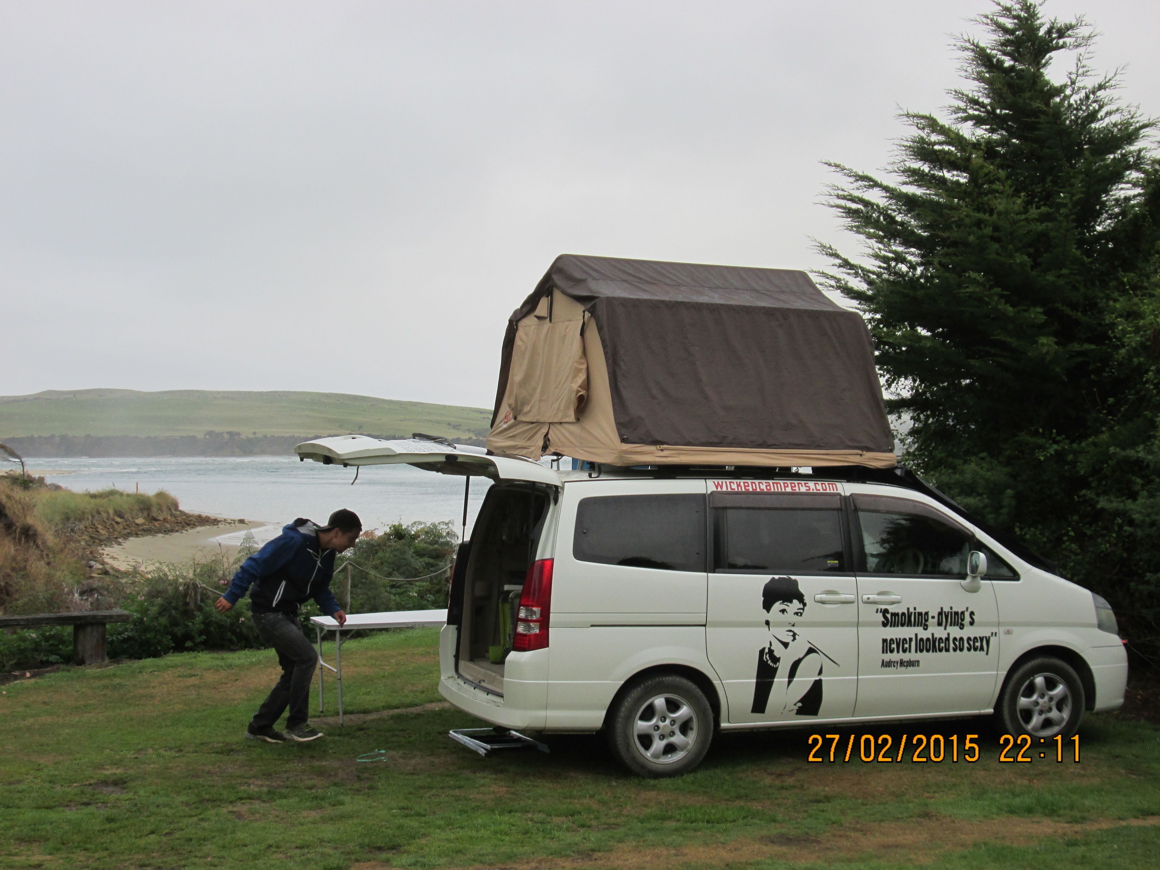 A white van with a tent on top, parked by a beach with a person nearby.