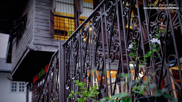 A wrought iron gate with intricate designs, leading to a building with wooden siding and a window with yellow-tinted glass. - Baan Chang Ton (Surin)