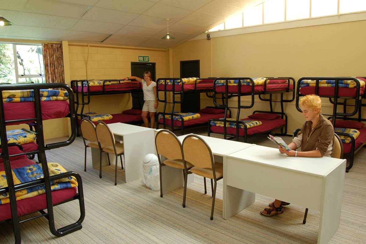 A dormitory room with bunk beds, a desk, chairs, and a trash bin.