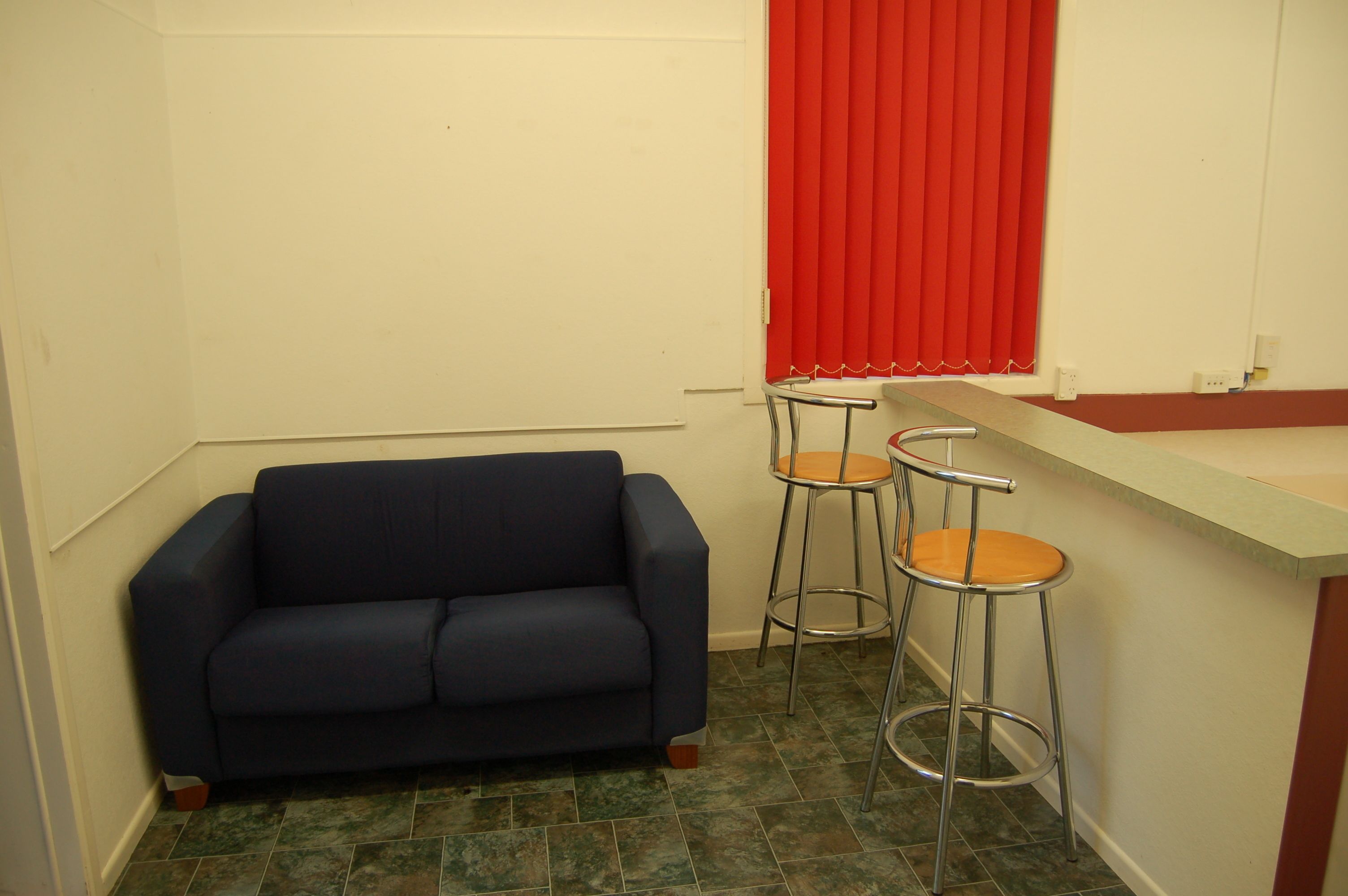 A small room with a dark blue sofa, two metal barstools, a red curtain, and a white wall.