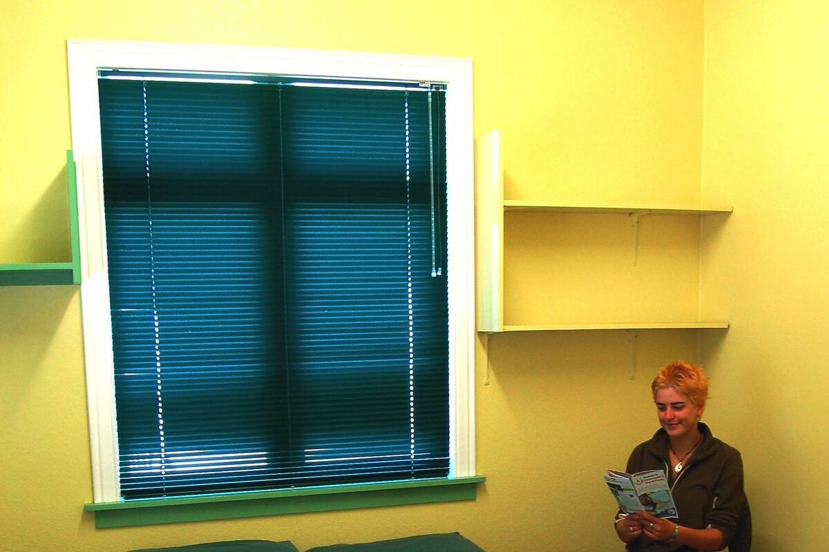 A room with a bed, a window with blinds, a shelf, and a person reading a book.