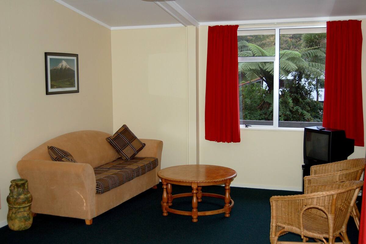 A living room with a beige sofa, a round wooden table, wicker chairs, a television, and a window with red curtains.