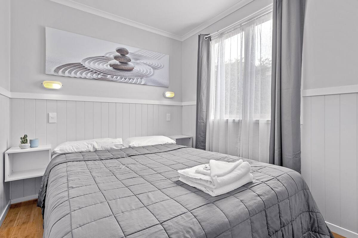 A neatly made bed with a grey quilt, a white pillow, and a folded towel on top. There is a wall-mounted artwork depicting a stack of stones.