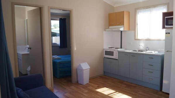 A compact kitchen with a microwave, stove, sink, and cabinets. There is a small trash bin and a window providing natural light.