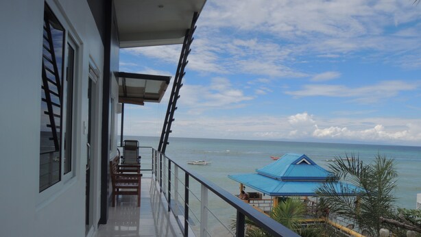 A balcony with a view of the sea, a blue-roofed structure, and a palm tree. - L&M Hearthstones Lodge (Oslob)