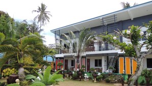 A modern building with a covered outdoor area, surrounded by lush tropical plants and palm trees. - L&M Hearthstones Lodge (Oslob)