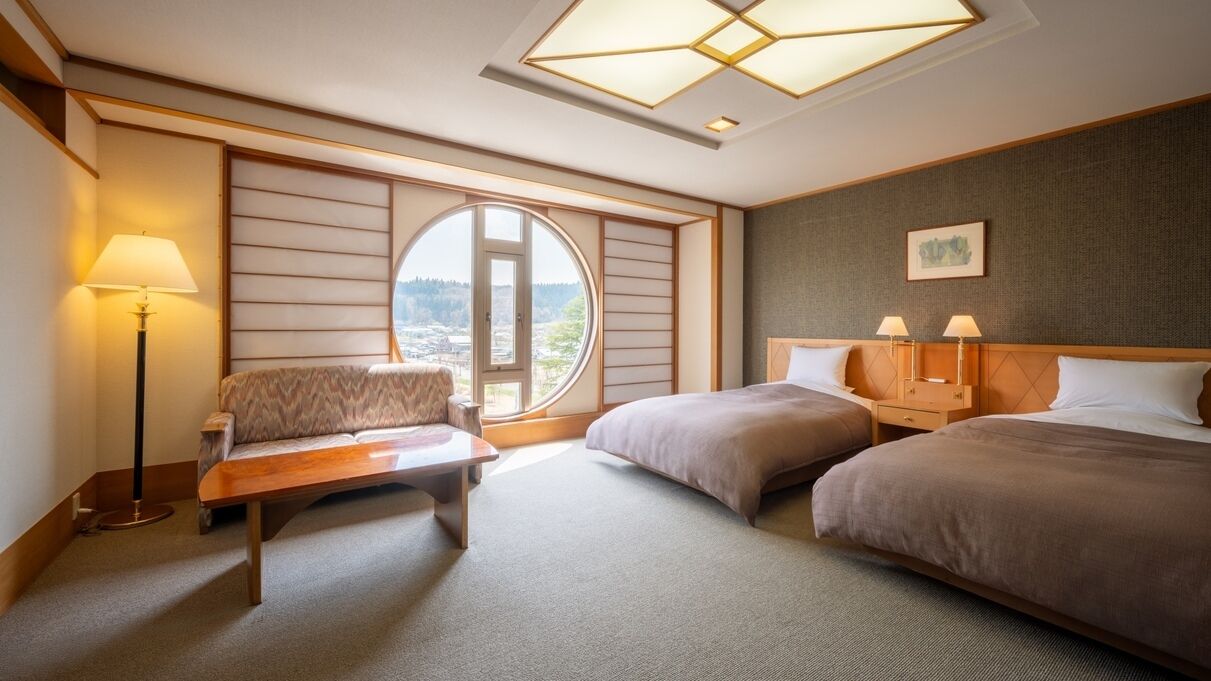 A hotel room with a sofa, two beds, a table, a lamp, a window with a view, and a ceiling light fixture.