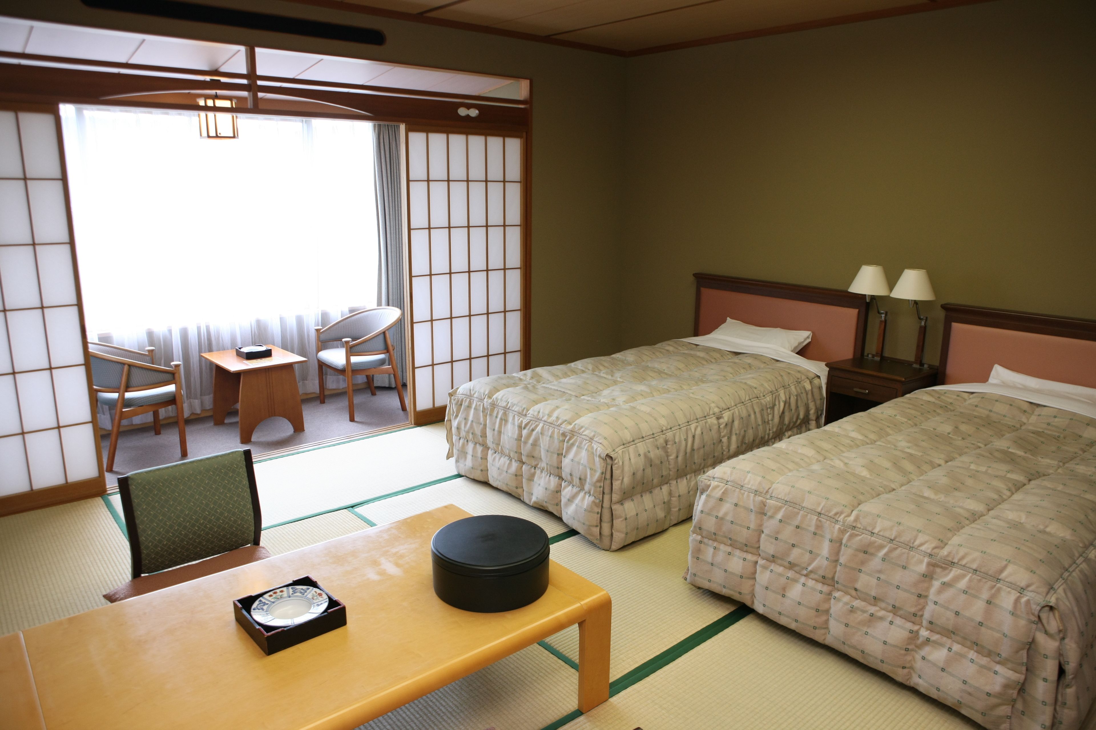 Habitación de hotel con suelo de tatami, dos camas, una mesita con juego de té, una silla y un divisor corredero shoji.
