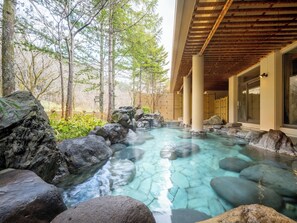 A hot spring area with clear water, surrounded by rocks and trees, and a building with large windows. - Hotel Kazuno (Kazuno)