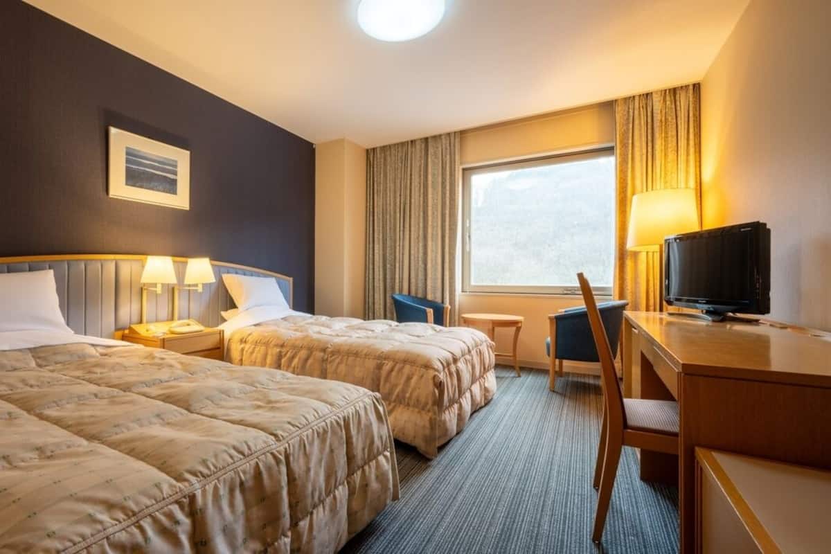 A hotel room with two beds, a desk, a chair, a TV, a window with curtains, and a framed picture on the wall.