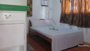 Una cama individual con colchón y almohadas blancos, cabecera de madera, un elemento decorativo verde sobre la cama, un aire acondicionado instalado en la pared y una ventana con cortinas marrones.