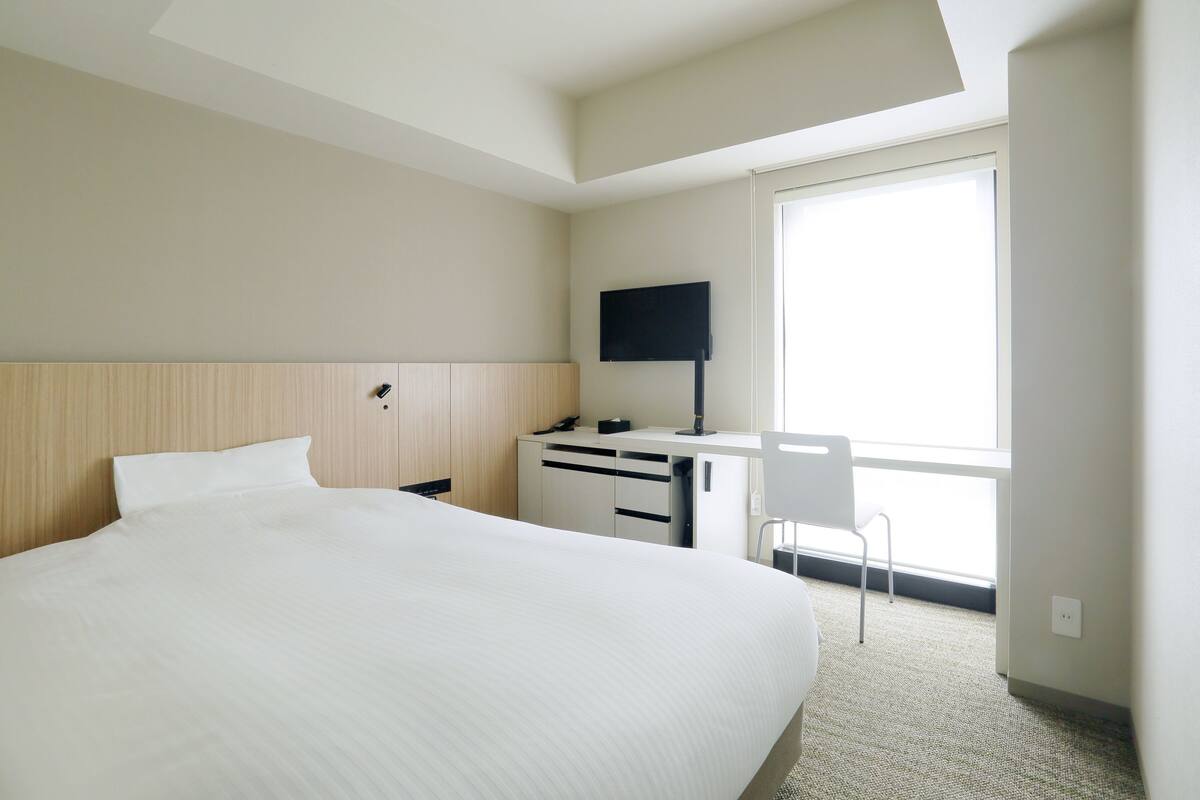 A hotel room with a large bed, a desk with a chair, a flat-screen TV, and a window providing a view.