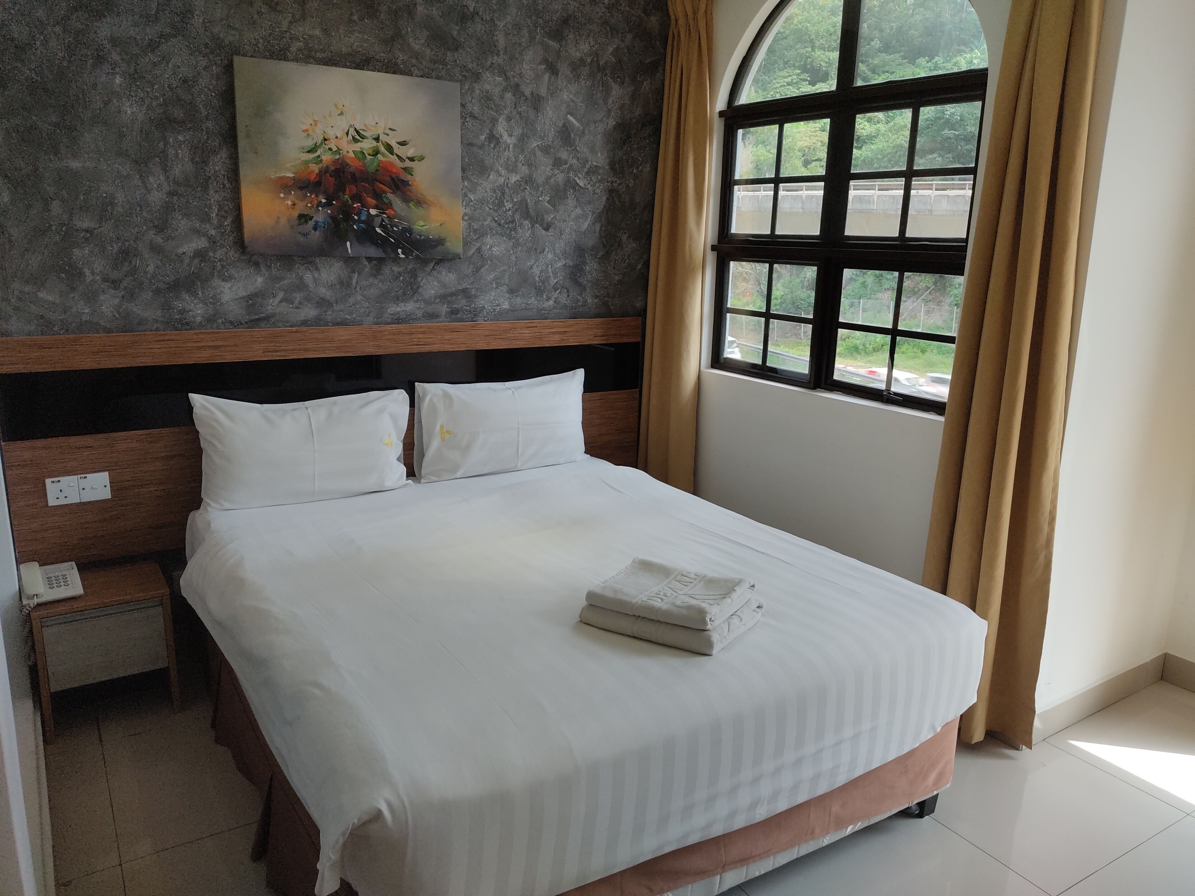 A neatly made bed with white linens and a folded towel, a wooden headboard, a painting of flowers on the wall, and a window with curtains.