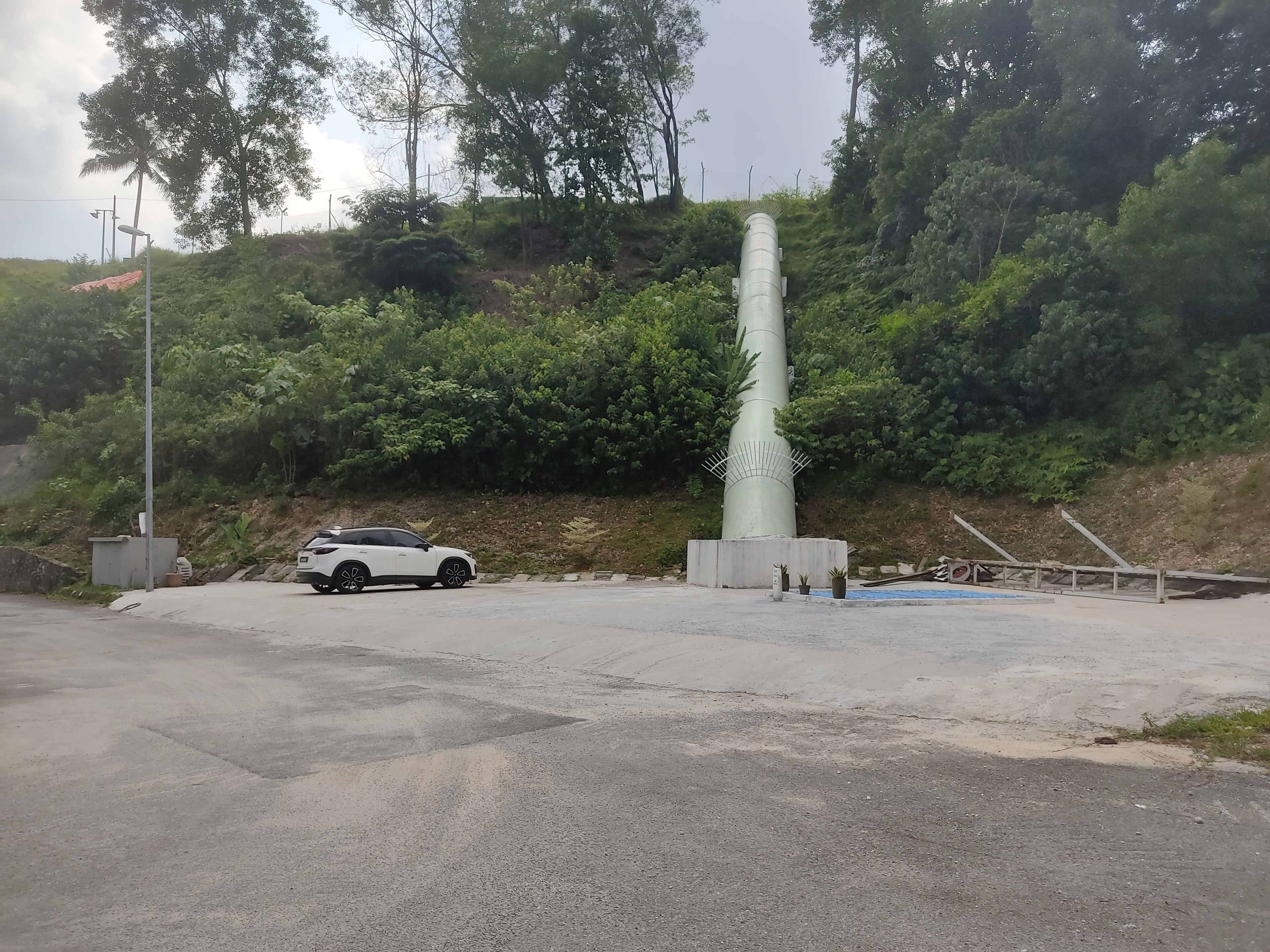 Ein weißes Auto ist in der Nähe einer großen, zylindrischen Struktur mit einem Metallzaun geparkt, umgeben von Grünflächen und einem Hügel.