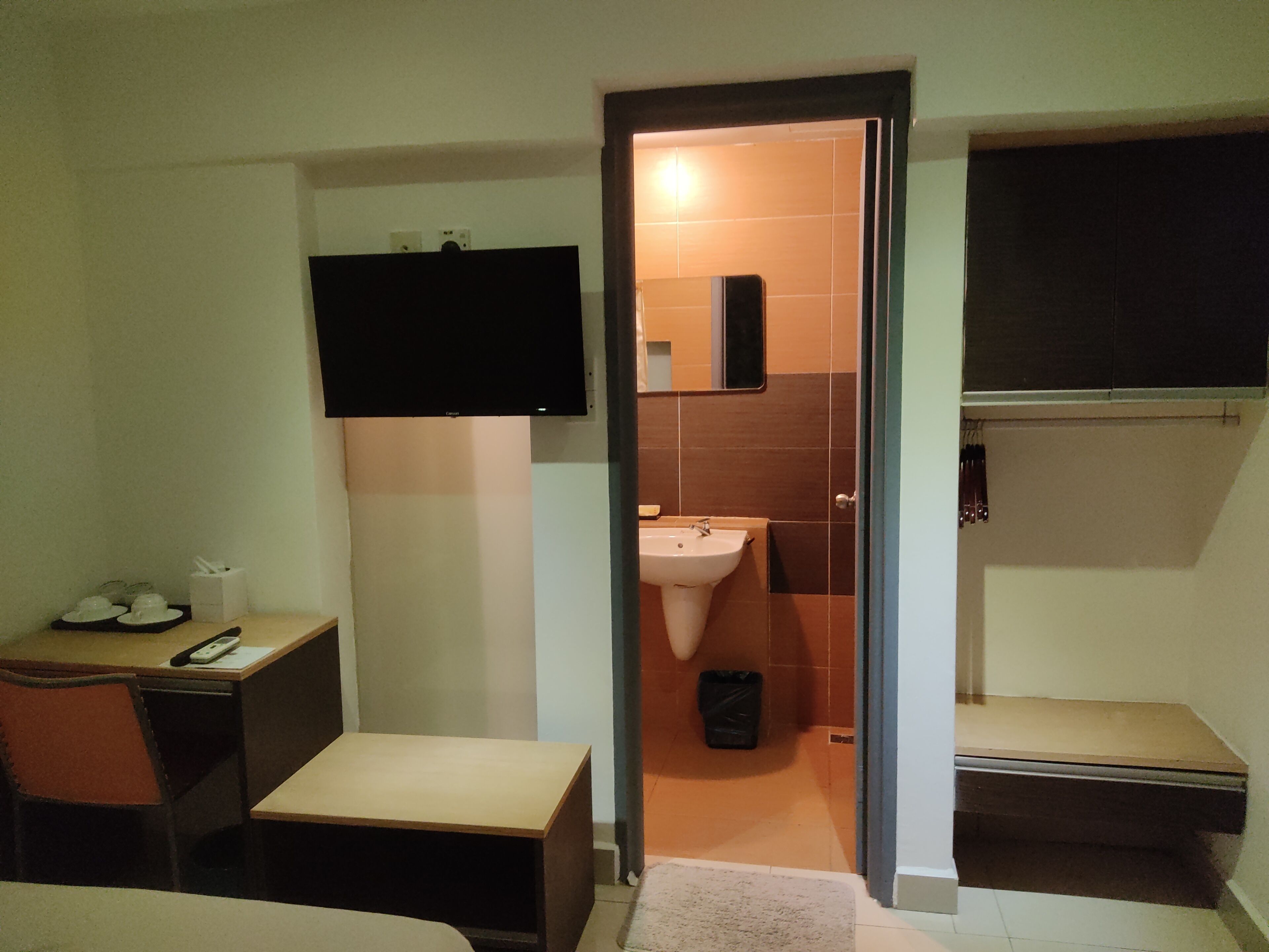 A compact hotel room with a bed, a desk with a chair, a TV mounted on the wall, a small sink, and a trash bin.