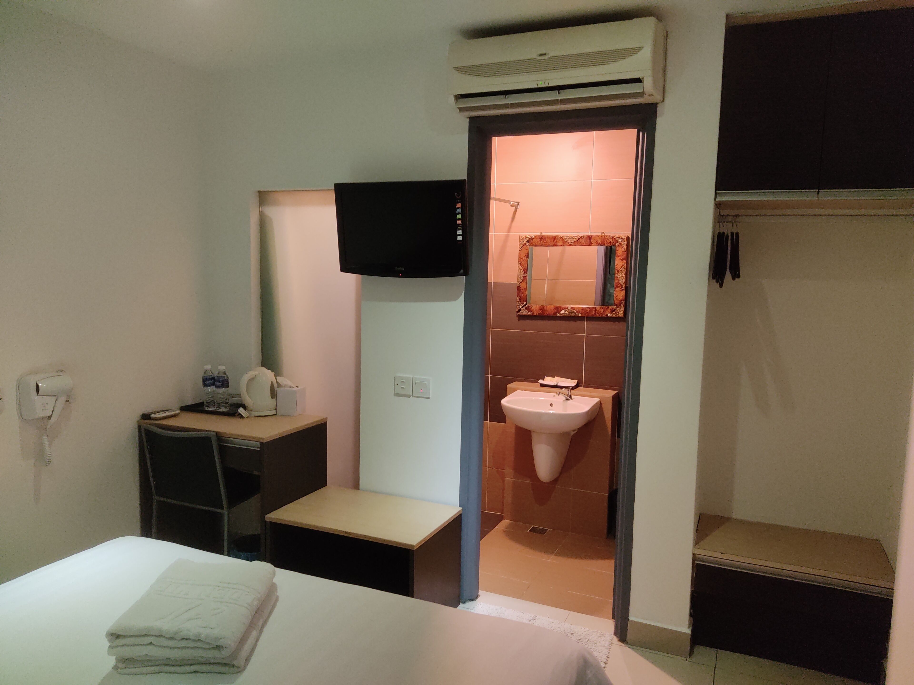 A hotel room with a bed, a small bedside table, a wall-mounted TV, a bathroom with a sink and mirror, and an air conditioning unit.