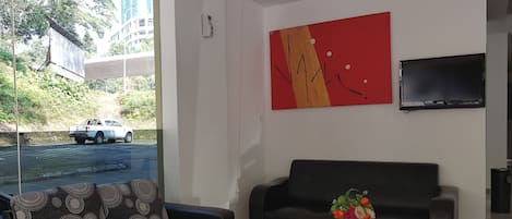 A modern living room with a black sofa, a coffee table with a vase of flowers, a red abstract painting, and a TV mounted on the wall.