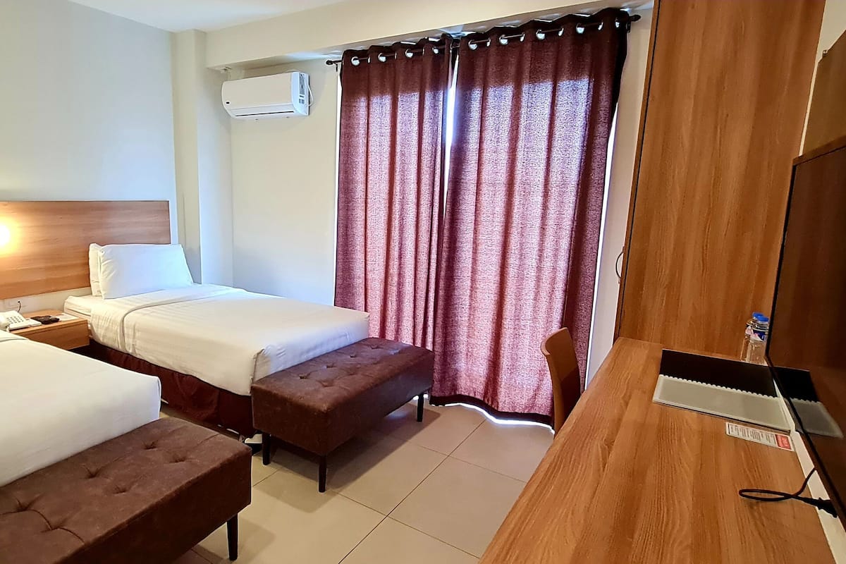 Quarto de hotel com duas camas, uma escrivaninha de madeira, televisão e janela com cortinas.