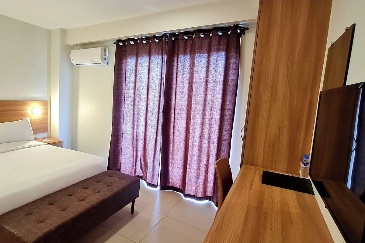 Quarto de hotel com cabeceira de madeira, cama com roupa de cama branca, mesa de cabeceira com telefone e carregador, um banco ao pé da cama, TV em suporte de madeira e janela com cortinas roxas.
