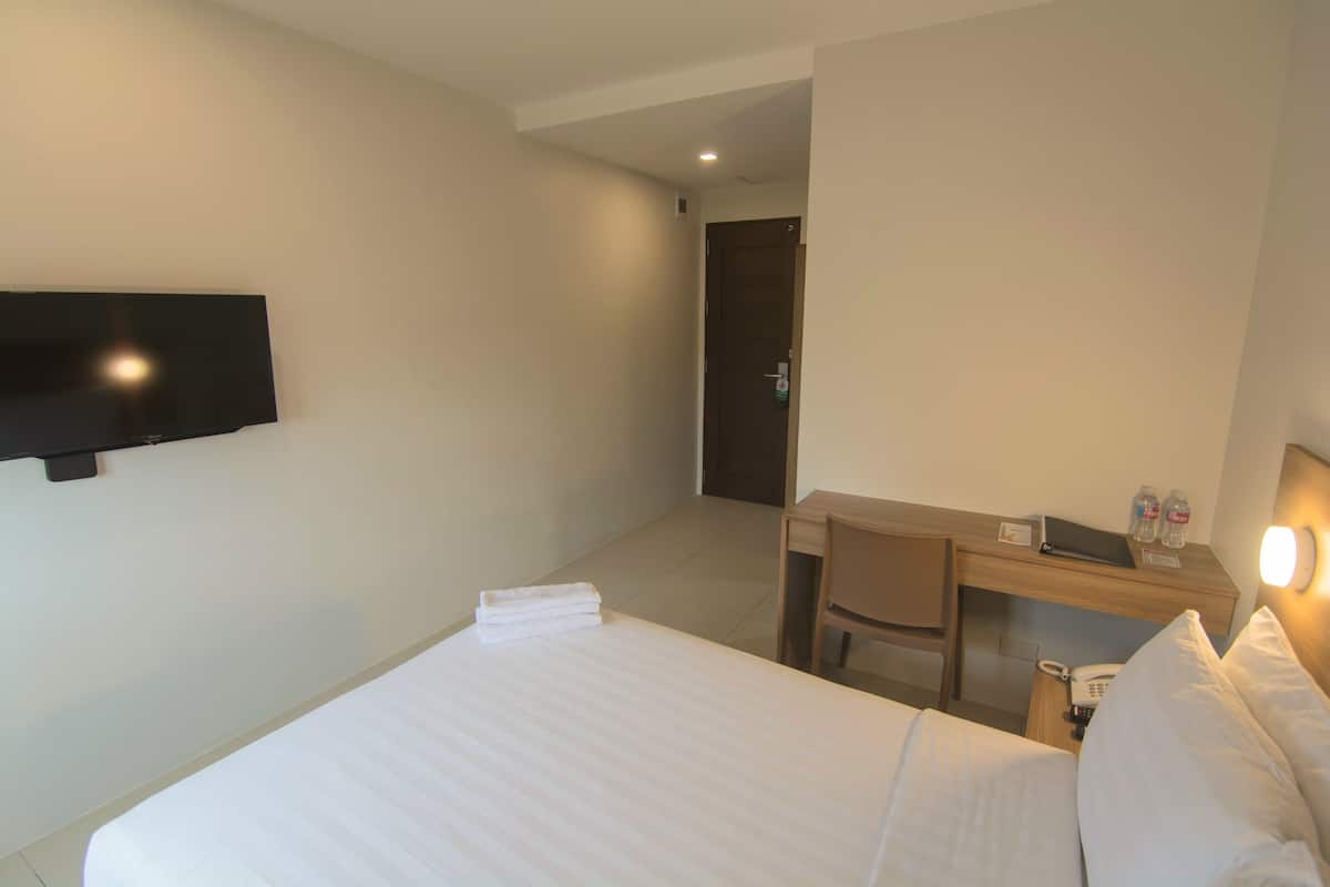 Quarto de hotel com cama, escrivaninha, cadeira, televisão de tela plana na parede e mesa de cabeceira com telefone e garrafa.