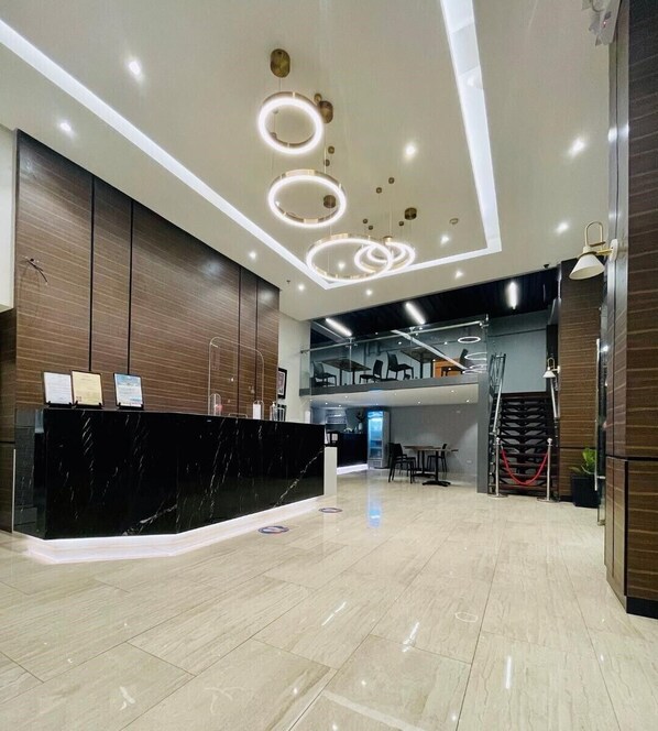 A modern hotel lobby with a marble reception desk, a staircase, and a mezzanine level with seating.