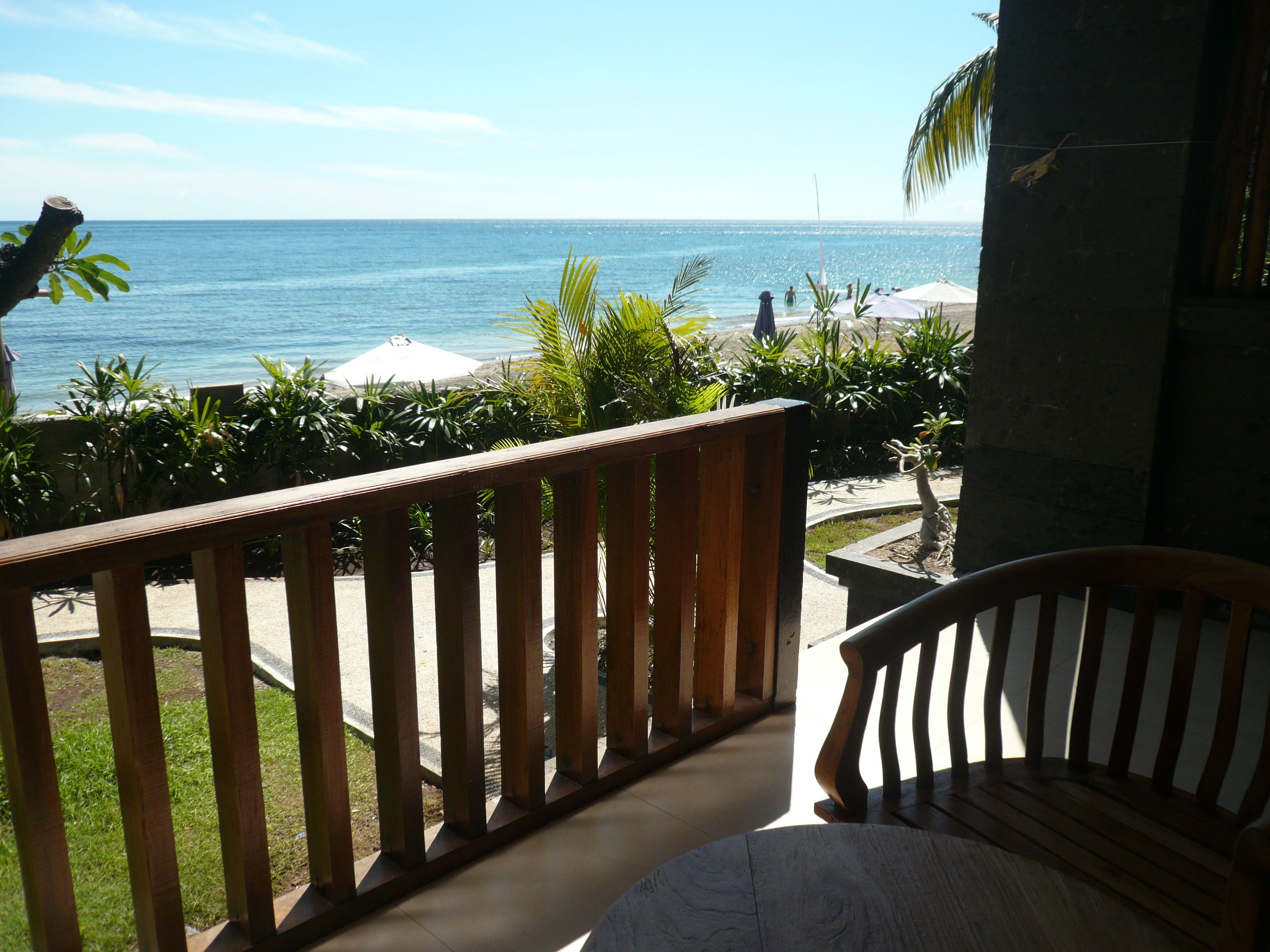 A wooden balcony with a view of the ocean, surrounded by greenery and umbrellas.