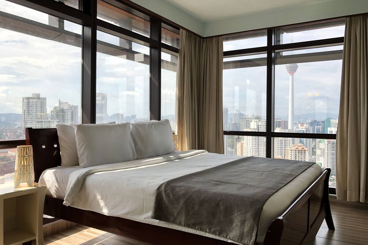 Une chambre d’hôtel moderne avec un grand lit, une table de chevet et une vue sur le paysage urbain grâce à de grandes fenêtres.