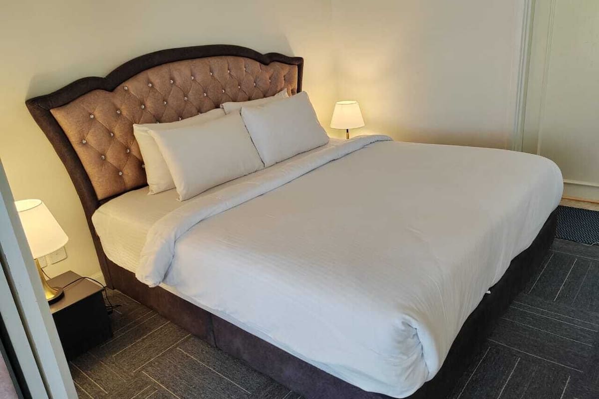Un lit bien fait, avec une tête de lit capitonnée, du linge de lit blanc et une table de chevet surmontée d’une lampe.