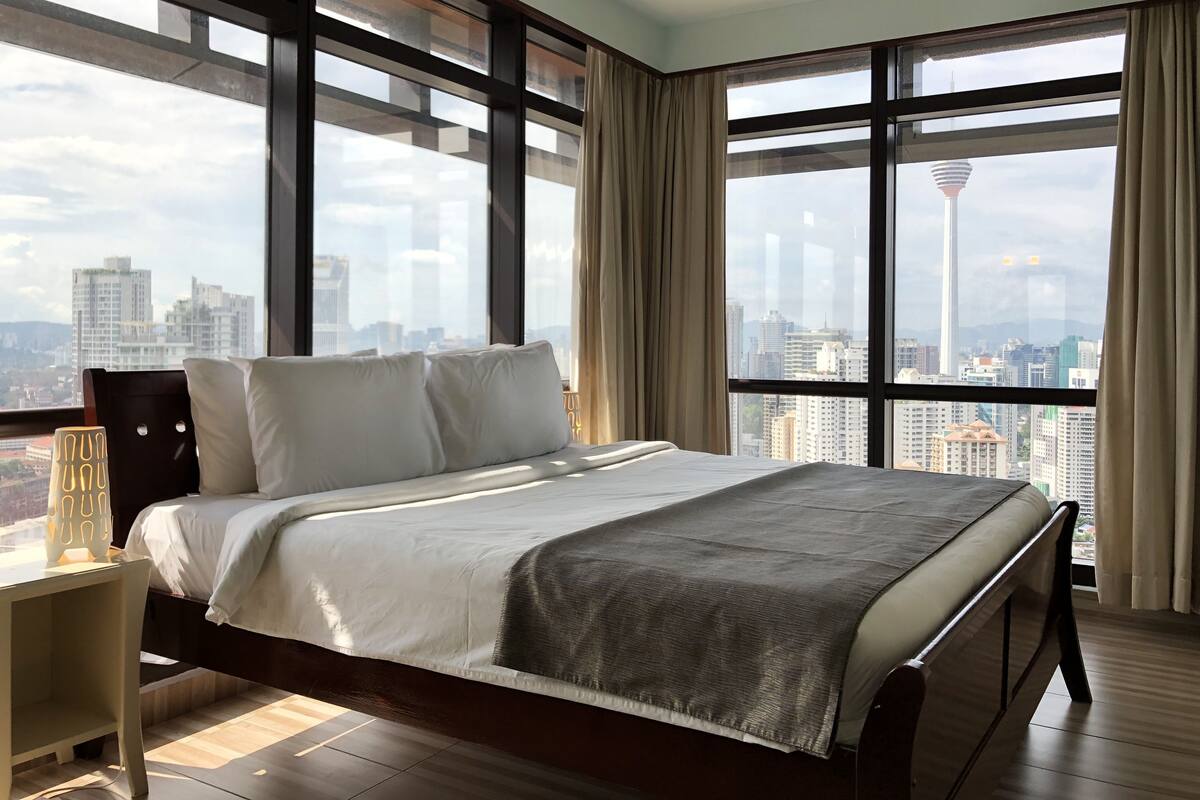 Une chambre d’hôtel moderne dotée d’un grand lit, d’une table de chevet et offrant une vue sur le paysage urbain grâce à de grandes fenêtres du sol au plafond.