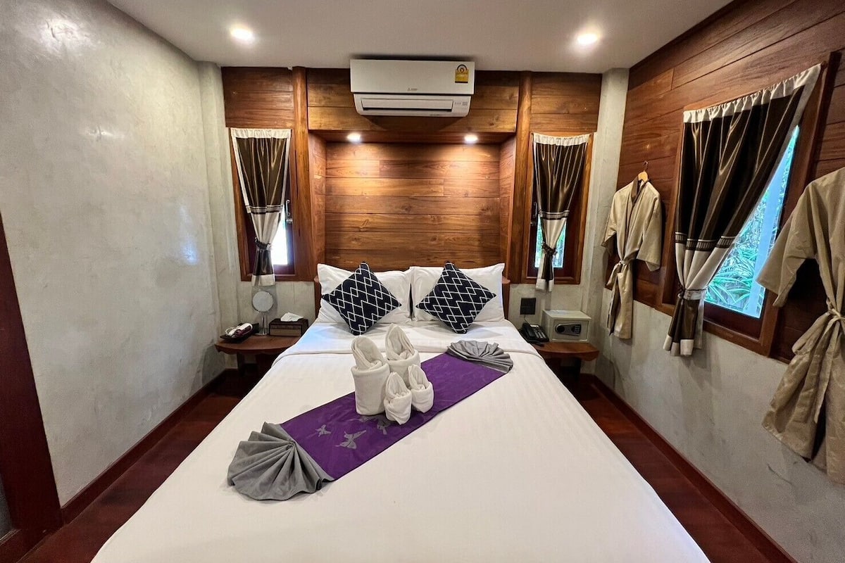 A hotel room with a large bed, wooden paneling on the walls, an air conditioning unit, a bedside table with a lamp, and a window with curtains.