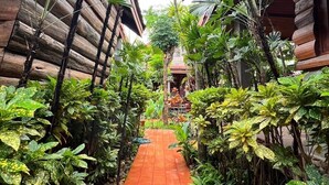 Un sendero estrecho bordeado por exuberante vegetación tropical y construcciones de madera, que conduce a una zona central con suelo de baldosas y un pequeño edificio.