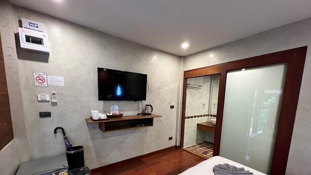 Habitación de hotel con cama, televisor montado en la pared, estante pequeño con artículos de aseo, cesto de basura y puerta corredera al baño.