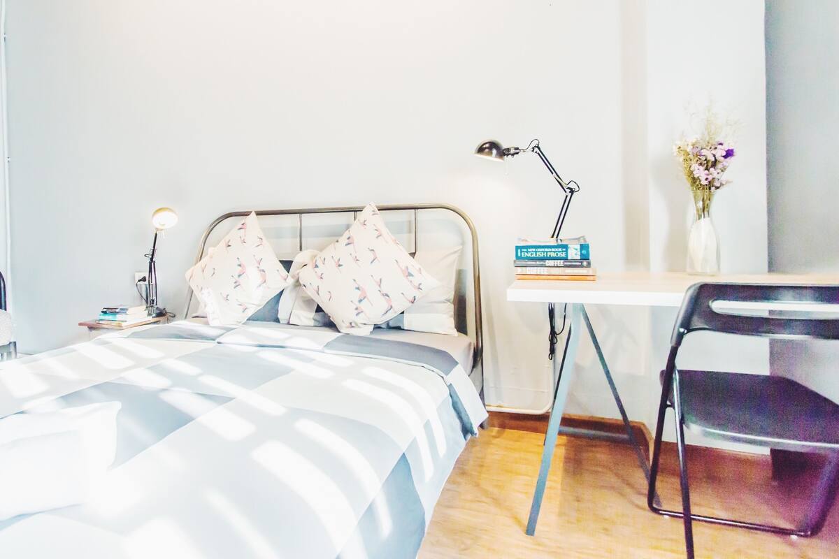 A neatly arranged bedroom with a bed, bedside table, lamp, books, and a desk with a chair.