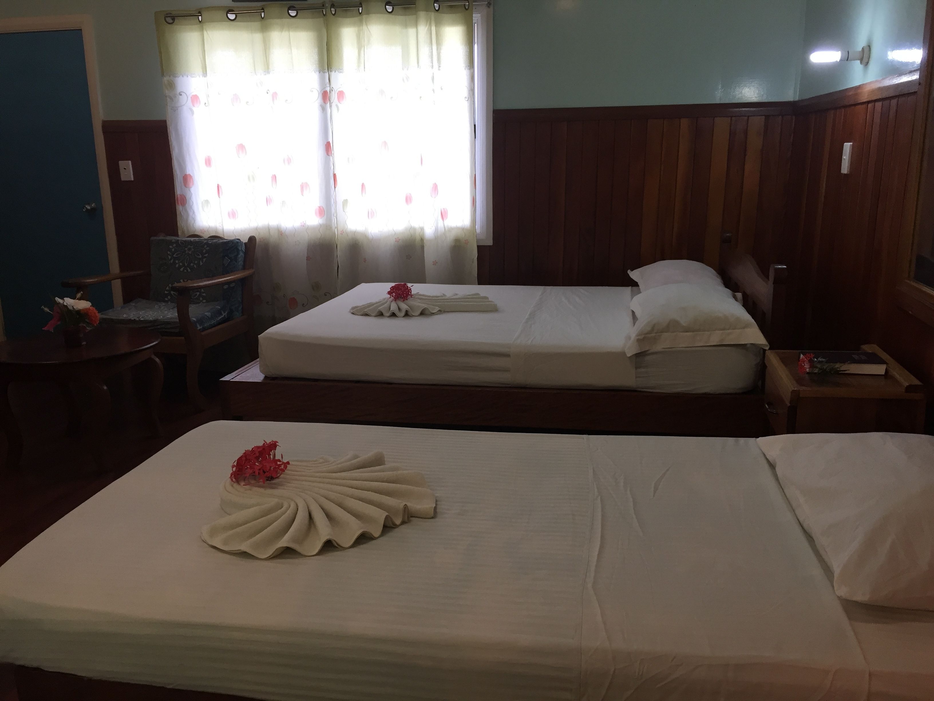 A hotel room with two beds, a wooden headboard, a small table with a book, a chair, and a window with curtains.