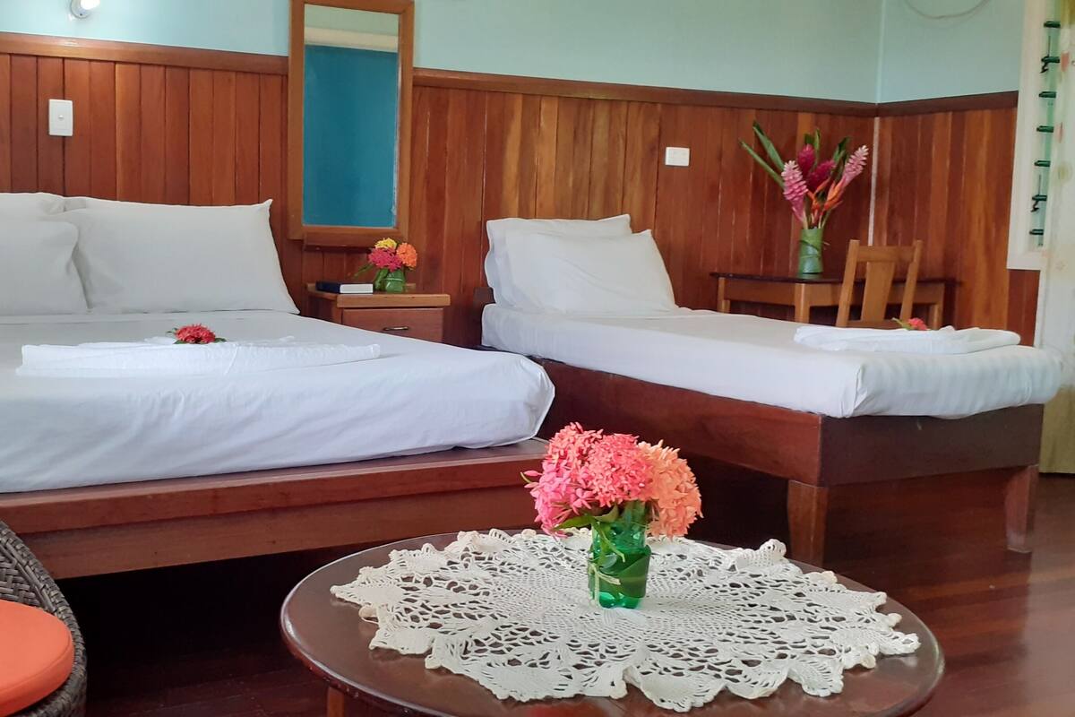 A hotel room with two beds, wooden headboards, a wicker chair, a small table with a lace tablecloth, and a vase with flowers.