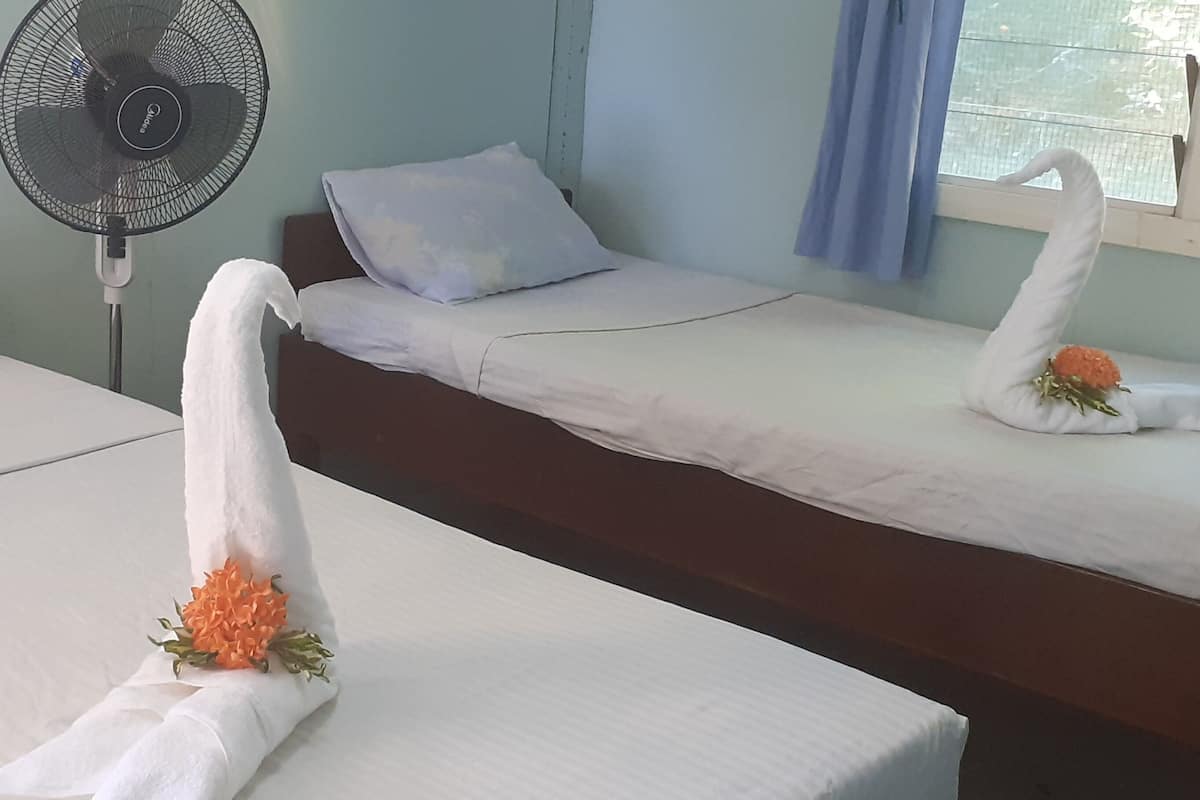 A hotel room with two beds, each with a white pillow and a decorative swan made of a towel with an orange flower. A standing fan is visible in the room.