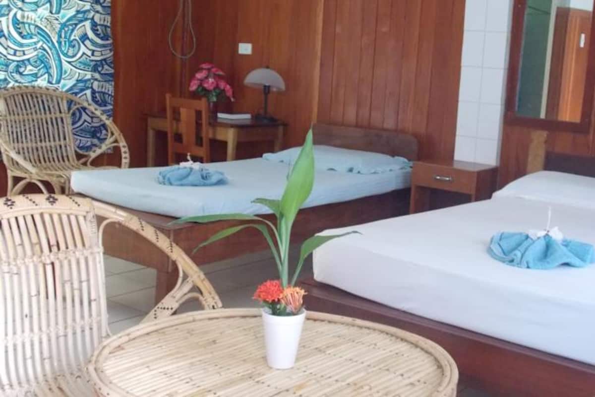 A room with two beds, a wicker chair, a small table with a lamp, and a potted plant.