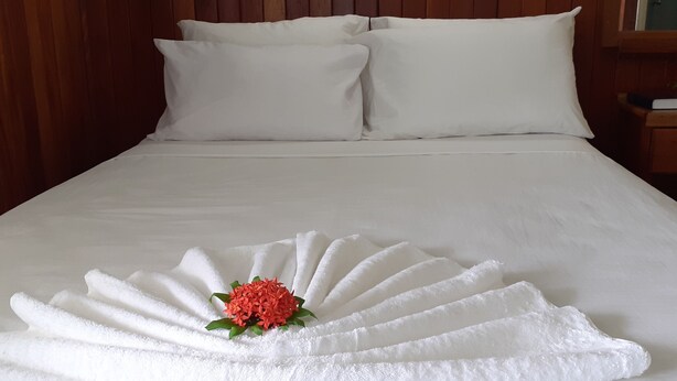A neatly made bed with white linens and a towel arrangement with a flower centerpiece. - Rekona Lodge (Gizo)