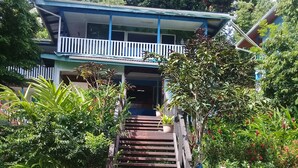 A house with a porch, a staircase leading to the entrance, and a garden with various plants. - Rekona Lodge (Gizo)