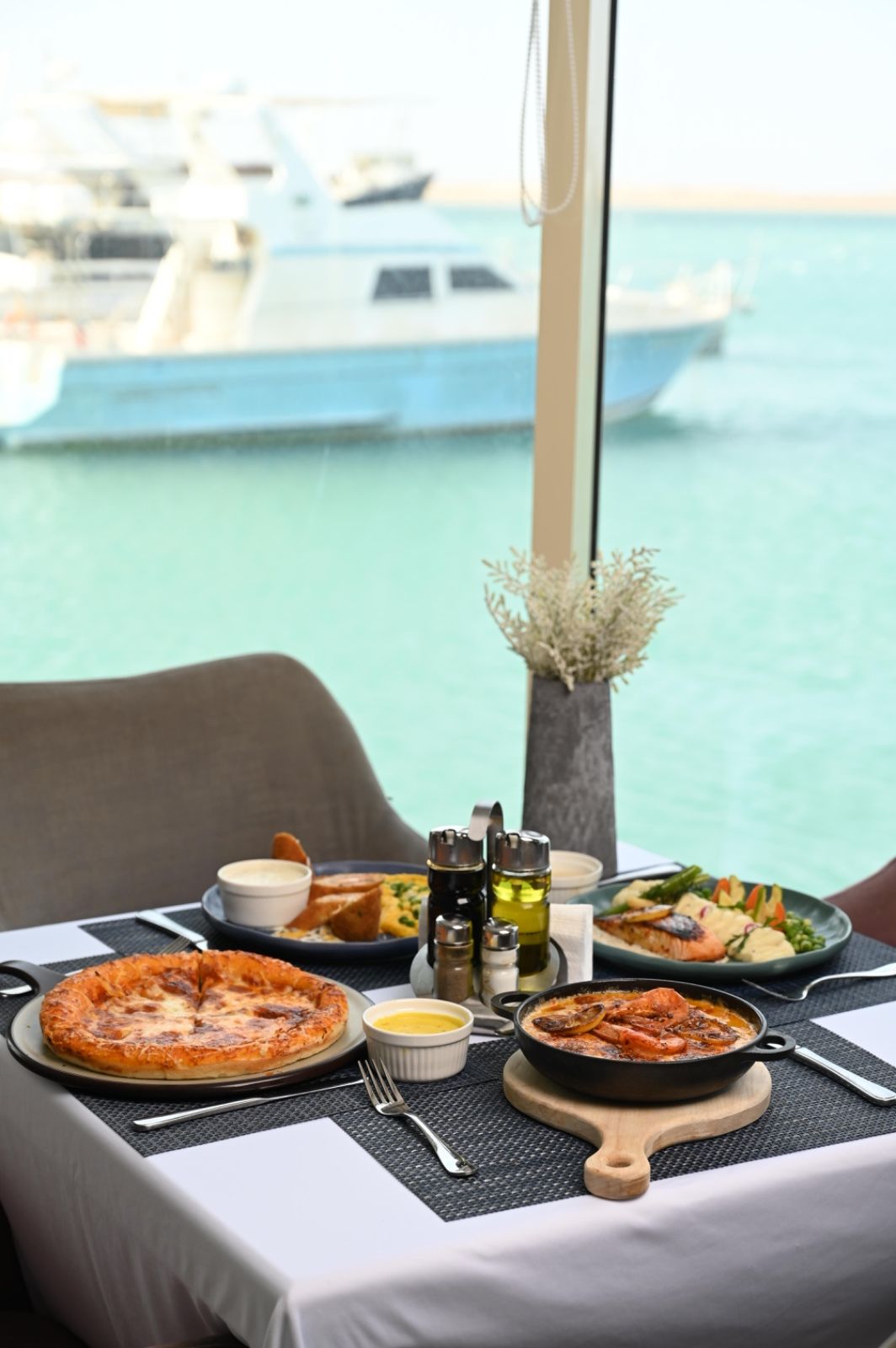 A table set with various dishes, including pizza and seafood, overlooking a marina with boats.