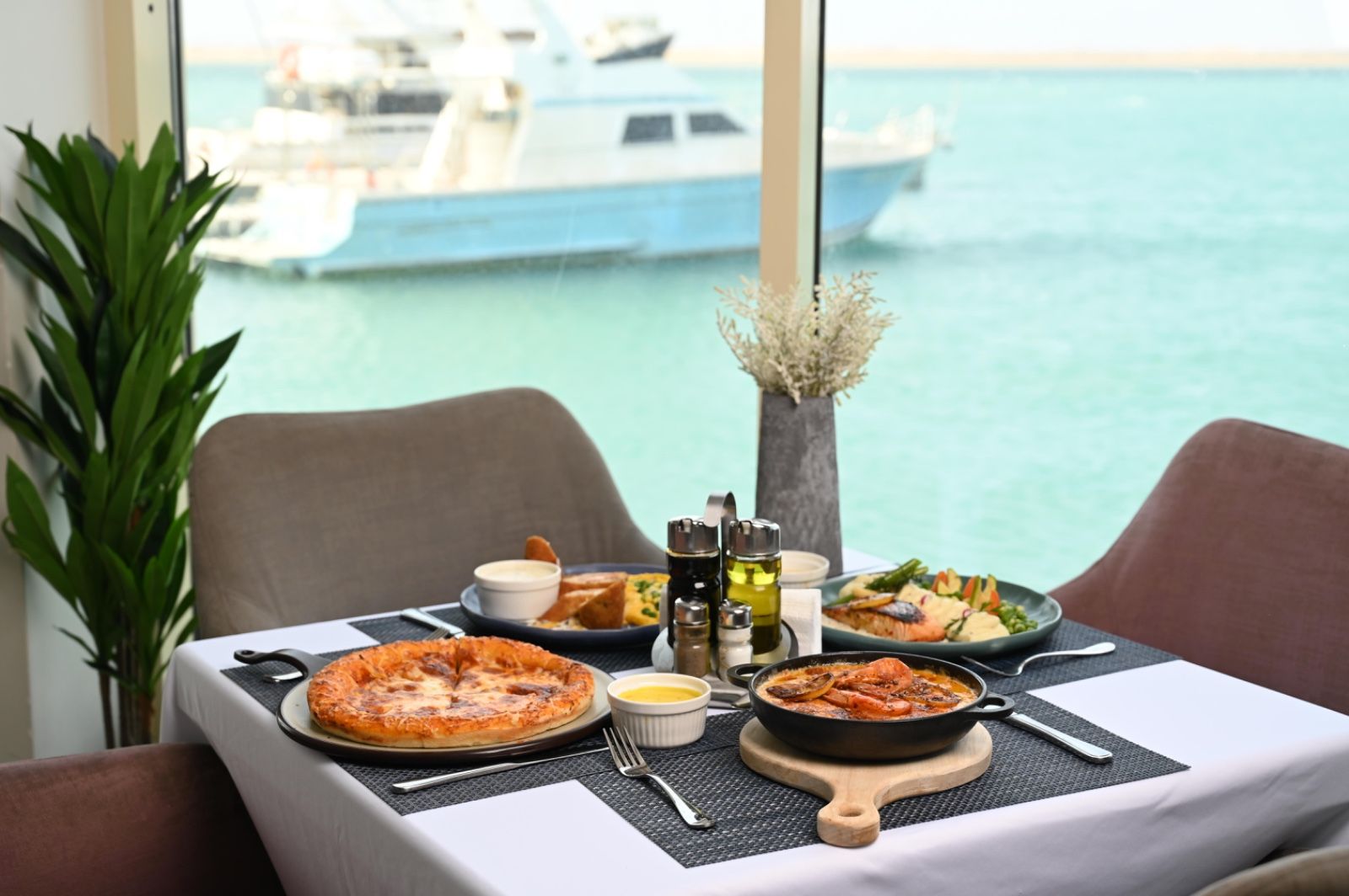 Una mesa de comedor con diversos platos, incluyendo pizza, un plato de mariscos y una ensalada, con un barco en el agua al fondo.