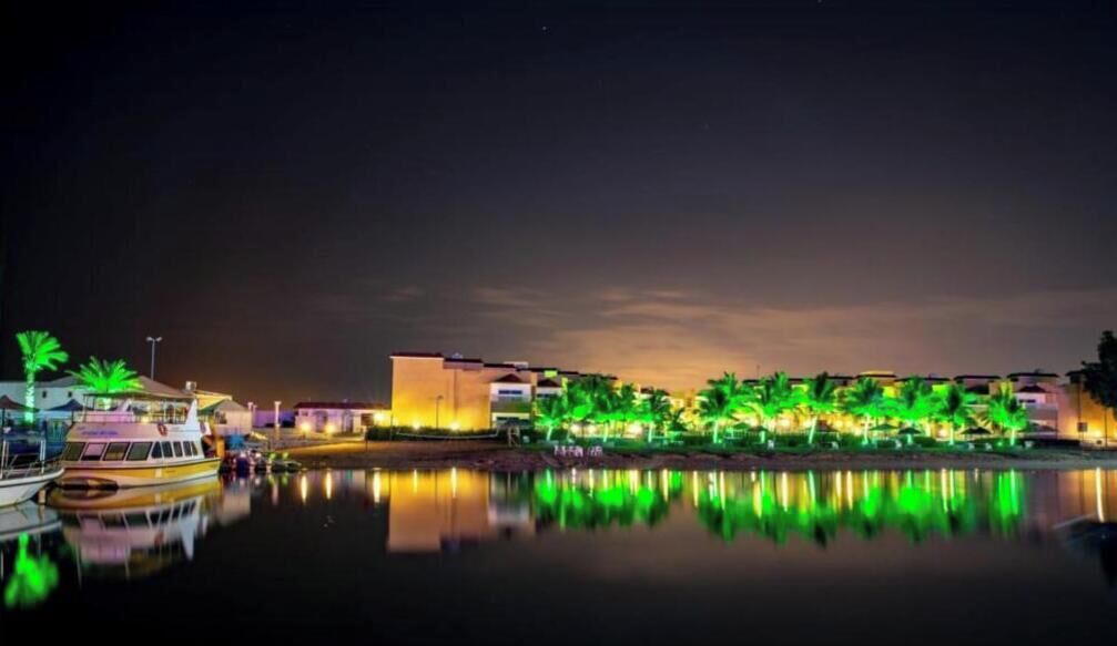 A waterfront hotel with a reflection in the water, a boat docked at the pier, and buildings with illuminated windows.