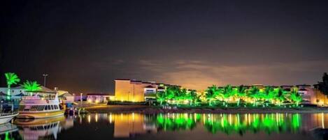 A waterfront hotel with a reflection in the water, a boat docked at the pier, and buildings with illuminated windows.