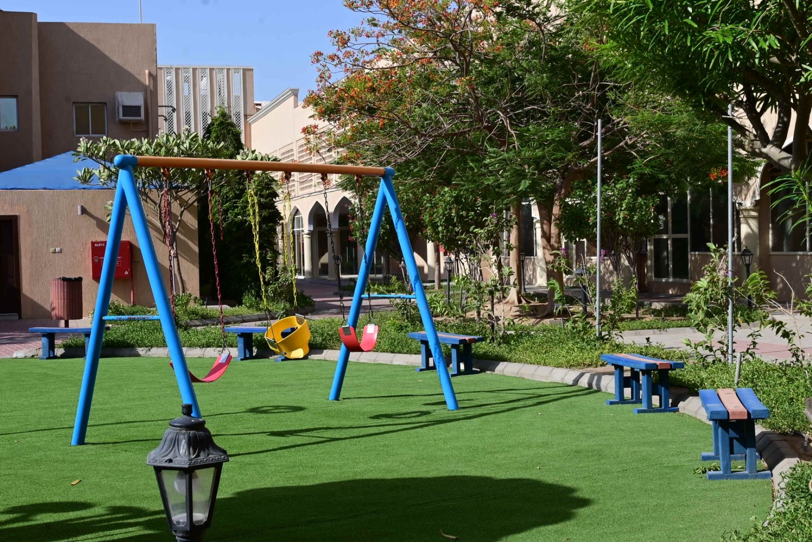 A playground with swings and benches, surrounded by greenery and buildings.